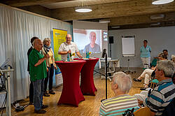 Ein Referent spricht vor einer Gruppe älterer Menschen, die an Tischen sitzen. Auf einer Leinwand ist eine Person per Videokonferenz zugeschaltet. Im Raum stehen Tische mit Materialien und Getränken.
