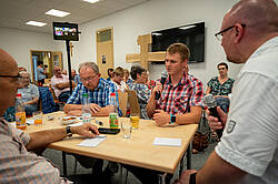 Ein Referent spricht vor einer Gruppe älterer Menschen, die an Tischen sitzen. Auf einer Leinwand ist eine Person per Videokonferenz zugeschaltet. Im Raum stehen Tische mit Materialien und Getränken.