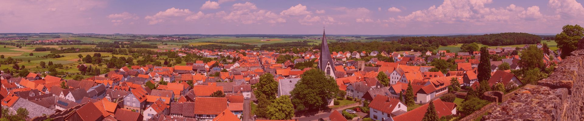 Panoramaaufnahme eines Dorfes mit vielen roten Dächern, einer Kirche mit spitzem Turm in der Mitte und Feldern im Hintergrund.
